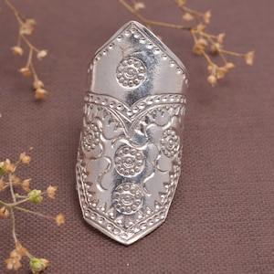 Puede incluir: Un anillo plateado con un diseño alargado. El anillo presenta intrincados patrones florales y circulares, con una parte superior puntiaguda y una banda ancha. El metal tiene un acabado pulido, que refleja la luz. El anillo probablemente está hecho de plata de ley o un metal similar.