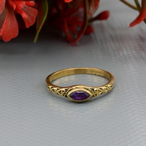 May include: A gold-colored ring with an oval, deep purple gemstone at the center. The band features an ornate, repeating pattern. The ring is set against a gray background, with red floral accents in the upper corners.