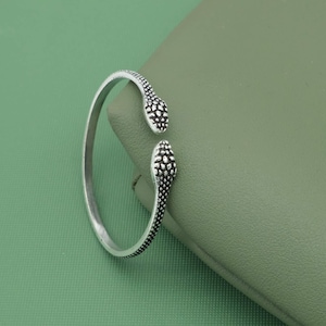 May include: A silver cuff bracelet with a snake design. The bracelet has two snake heads at the ends, with textured detailing. The bracelet is displayed on a green surface.