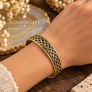 May include: A gold-toned metal cuff bracelet featuring an intricate, continuous Celtic knot pattern engraved along the outer surface. The bracelet has a polished finish with darkened recessed areas that highlight the woven design details.