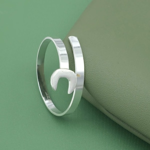 Könnte beinhalten: Ein silberfarbener Ring mit einem halbmondförmigen Design. Der Ring umschließt den Finger mit einer glatten, polierten Oberfläche. Der Ring wird auf einer grünen Oberfläche präsentiert.