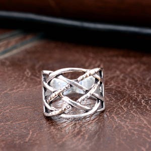 Puede incluir: Un anillo plateado con un intrincado diseño de nudo celta. El anillo presenta bandas entrelazadas que crean un patrón calado texturizado. El anillo está sobre una superficie marrón similar al cuero.