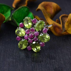 May include: A silver ring featuring a cluster of oval-cut peridot gemstones in a light green hue, interspersed with smaller, round, deep pink ruby gemstones. The ring's design is floral, with the gemstones arranged in a circular pattern.
