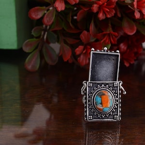May include: A silver-toned ring with an open rectangular top and an oval-shaped turquoise and orange stone. The ring has a decorative, ornate design and is set against a dark wooden surface with red flowers in the background.