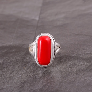 May include: A silver ring featuring a vibrant red, rectangular gemstone. The gemstone is set within an oval frame, and the ring has a split band design. The ring is displayed against a gray background.