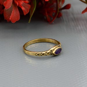 May include: A gold-colored ring with an oval, purple gemstone. The ring has decorative filigree patterns along the band and around the gemstone setting. The ring is set against a gray background with red flowers in the upper corner.