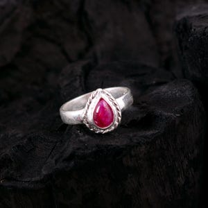 May include: A silver ring with a teardrop-shaped, deep red gemstone. The gemstone is set in a decorative bezel with a twisted rope design. The ring is displayed against a dark, textured background.
