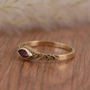 Könnte beinhalten: Ein goldfarbener Ring mit einem marquiseförmigen, tiefroten Edelstein. Der Ring hat ein verziertes Filigranmuster entlang des Rings und um die Edelsteinfassung. Der Ring wird auf einer Holzoberfläche präsentiert.