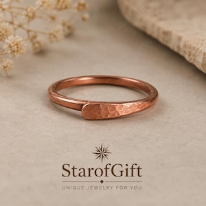 May include: A copper ring featuring a smooth, thin band that transitions into a wider, flattened section with a textured, hammered finish. The metal has a warm, reddish-brown metallic tone, and the design is simple and circular in shape.