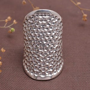 Puede incluir: Un dedal plateado con una superficie texturizada y con hoyuelos. El dedal es de forma cilíndrica y parece estar hecho de metal. El diseño es consistente en toda la superficie.