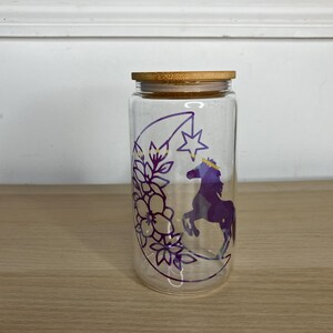 May include: A clear glass jar with a bamboo lid features a purple and gold design of a crescent moon, a star, flowers, and a unicorn.