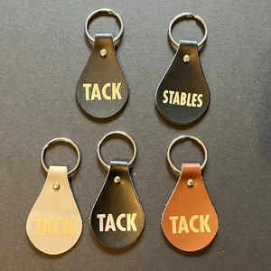Puede incluir: Varios llaveros de cuero en negro, marrón y beige. Cada uno tiene un llavero plateado y un accesorio remachado. Los llaveros están impresos con las palabras "TACK" y "STABLES" en color dorado. Diseñados para uso ecuestre.