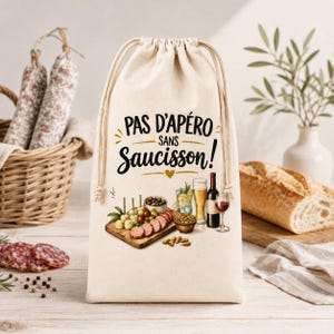 Peut inclure: Un sac en tissu beige à cordon de serrage avec le texte noir "PAS D'APÉRO sans Saucisson!" au-dessus d'une illustration colorée d'une planche à découper en bois avec du saucisson tranché, des olives, une bière, une bouteille et un verre de vin.
