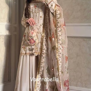 Pode incluir: Conjunto bordado creme com top de manga comprida, calças de perna larga e um lenço transparente. A roupa é adornada com detalhes florais e dourados. O texto "Vastrabella" é visível.