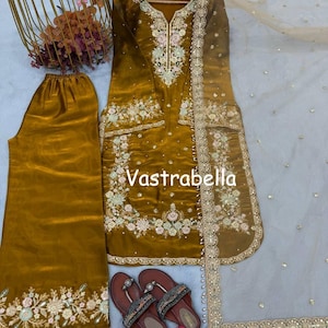 May include: A mustard yellow three-piece outfit featuring a long-sleeved tunic with intricate floral embroidery, matching wide-leg trousers with embroidered hems, and a sheer beige dupatta with a decorative border, labeled with the text Vastrabella.