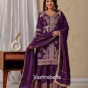 Könnte beinhalten: Ein tiefviolettes dreiteiliges Set bestehend aus einer langärmeligen Tunika, einem ausgestellten Rock und einem passenden Dupatta. Tunika und Rock sind mit silbernen Stickereien, Blumenmustern und Spiegeln verziert. Das Wort Vastrabella ist sichtbar.