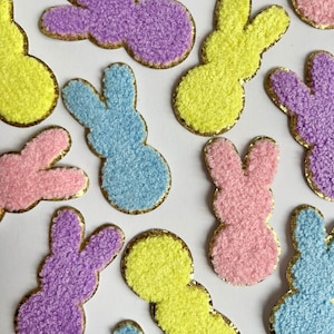 Könnte beinhalten: Eine Auswahl an bunten, hasenförmigen Aufnähern in Pastelltönen wie Gelb, Lila, Rosa und Blau. Jeder Aufnäher hat eine flauschige Textur und ist mit goldenem Glitzer umrandet. Ideal für Osterbastelarbeiten.