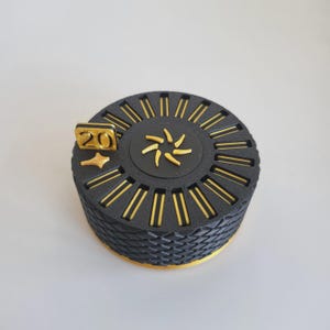 May include: A circular black container with a textured, woven-patterned side and a gold-coloured base. The top features a central gold star-like emblem, a gold number 20, and a small gold star, surrounded by a ring of gold-filled rectangular slots.