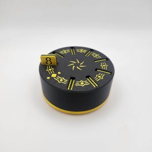 May include: A black and yellow circular game piece with a number 8 on a small tab. The top surface has a yellow starburst design and other yellow geometric patterns. The sides are black with a yellow trim.