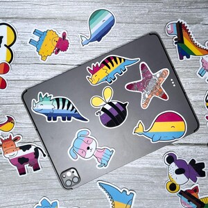 May include: A set of colorful stickers featuring various animals, insects, and sea creatures. The stickers are designed with different pride flags, including the rainbow, transgender, bisexual, and pansexual flags. The stickers are arranged on a gray tablet and a wooden surface.