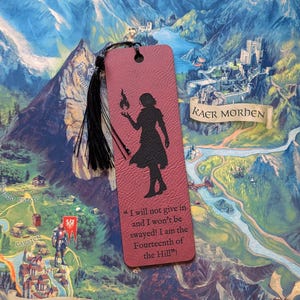 Peut inclure: Un signet bordeaux avec une silhouette noire d'une personne tenant une flamme. Le signet a un pompon noir et la citation, "Je ne renoncerai pas et je ne serai pas influencé ! Je suis le Quatorzième de la Colline !" sur lui. Le signet est sur une carte.