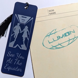 May include: A navy blue bookmark with a black tassel. The bookmark features a silver graphic of a couple and the text "See You At The Equator." A paper with the word "LUMON" is next to the bookmark.