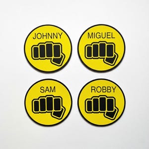May include: Four yellow circular coasters with black borders. Each coaster features a black fist graphic and a name: Johnny, Miguel, Sam, and Robby. The coasters are likely made of a durable material.