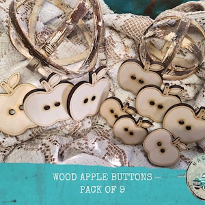May include: A collection of nine wooden apple-shaped buttons with two holes each. The buttons are light brown with a darker brown outline and stem. The text "WOOD APPLE BUTTONS - PACK OF 9" is visible at the bottom of the image.