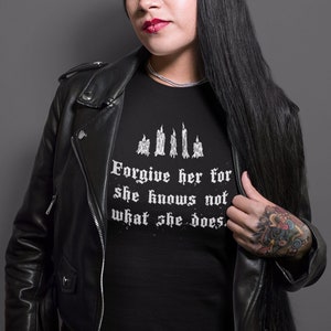 May include: A woman with long black hair is wearing a black leather jacket and a black t-shirt with the text "Forgive her for she knows not what she does." in white lettering.
