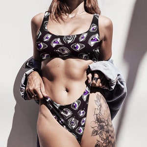May include: Black bikini set with an all-over eye print in white and purple. The top is a scoop neck style, and the bottoms are high-cut. The model is wearing a denim jacket. The background is a white wall with shadows.