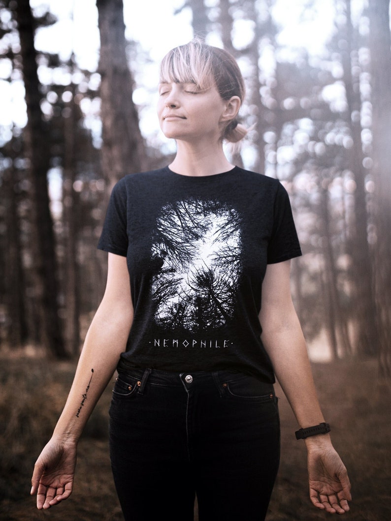 May include: A woman wearing a black t-shirt with a white graphic of trees and the text "NEMOPHILE." She is wearing black jeans and has a tattoo on her arm.