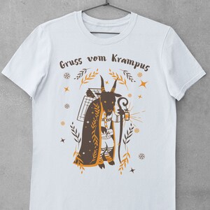 Puede incluir: Camiseta blanca con una ilustraci&oacute;n marr&oacute;n y naranja de una figura de Krampus sosteniendo una linterna y un bast&oacute;n. El texto "Gruss vom Krampus" est&aacute; impreso en la camiseta.