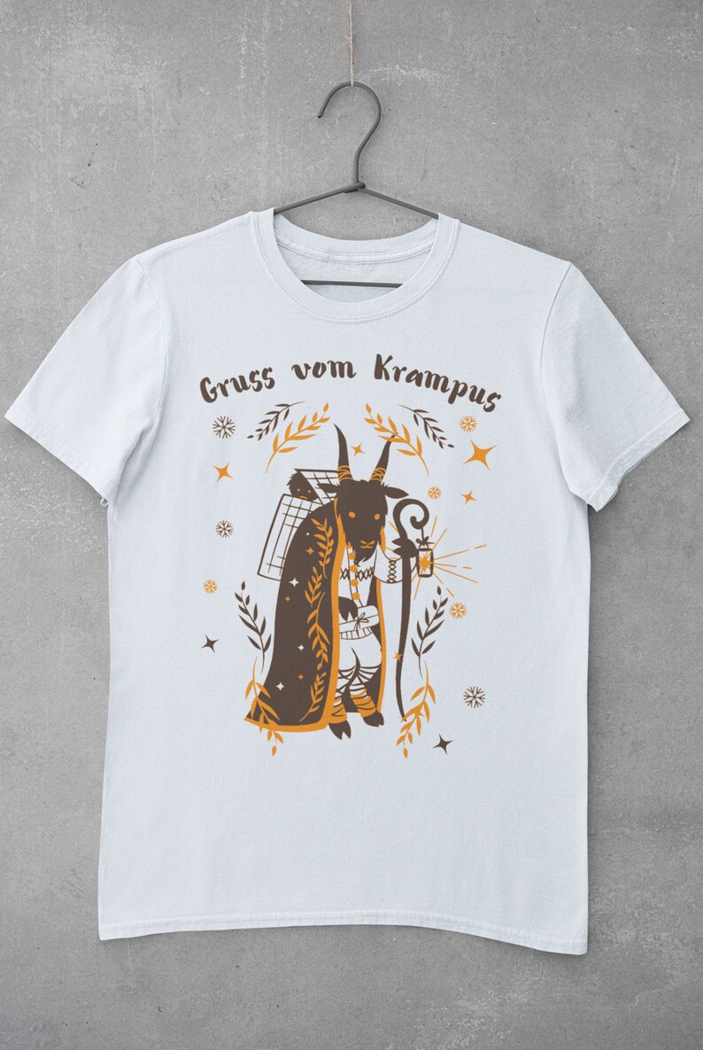 Puede incluir: Camiseta blanca con una ilustraci&oacute;n marr&oacute;n y naranja de una figura de Krampus sosteniendo una linterna y un bast&oacute;n. El texto "Gruss vom Krampus" est&aacute; impreso en la camiseta.