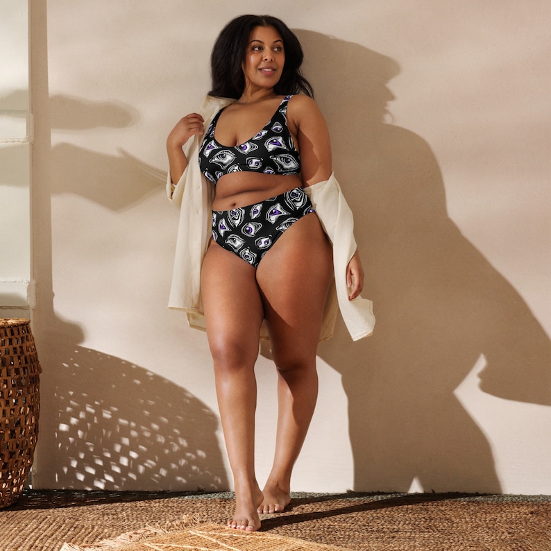 May include: Black two-piece swimsuit with a repeating eye pattern in white and purple. The top is a scoop-neck style, and the bottoms are high-waisted. The swimsuit is worn by a person with dark hair and skin, who is also wearing a light-colored, open shirt.