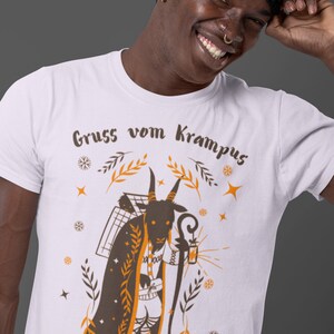 Puede incluir: Camiseta blanca con una ilustraci&oacute;n marr&oacute;n y naranja de una figura de Krampus. El Krampus es una criatura parecida a una cabra con cuernos, barba y una linterna. El texto "Gruss vom Krampus" est&aacute; impreso en la camiseta.