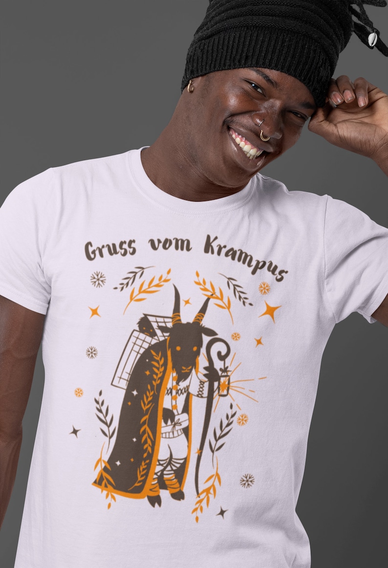 Puede incluir: Camiseta blanca con una ilustraci&oacute;n marr&oacute;n y naranja de una figura de Krampus. El Krampus es una criatura parecida a una cabra con cuernos, barba y una linterna. El texto "Gruss vom Krampus" est&aacute; impreso en la camiseta.
