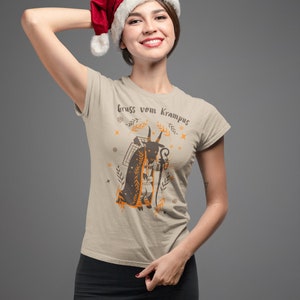 Puede incluir: Una mujer con una camiseta beige con un gr&aacute;fico de una figura de Krampus y el texto "gruss vom Krampus". Lleva un gorro de Pap&aacute; Noel rojo y una falda negra.