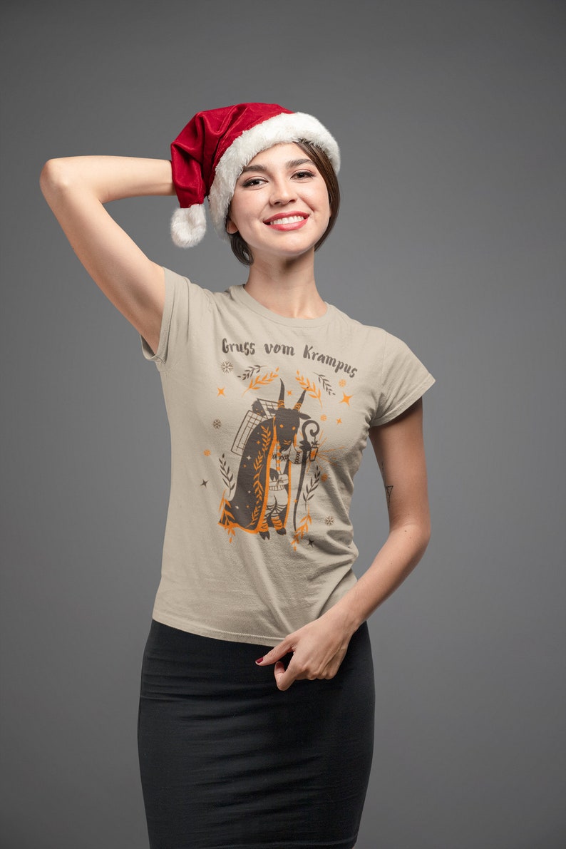 Puede incluir: Una mujer con una camiseta beige con un gr&aacute;fico de una figura de Krampus y el texto "gruss vom Krampus". Lleva un gorro de Pap&aacute; Noel rojo y una falda negra.