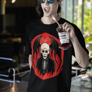 K&ouml;nnte beinhalten: Ein schwarzes T-Shirt mit einem Aufdruck eines Vampirs mit roten Augen und Fl&uuml;geln. Der Vampir tr&auml;gt einen schwarzen Anzug und hat ein wei&szlig;es, sch&auml;delartiges Gesicht. Der Aufdruck ist von einem roten Kreis mit Flammen umgeben. Die Person, die das Shirt tr&auml;gt, h&auml;lt einen Plastikbeutel mit roter Fl&uuml;ssigkeit und einem Strohhalm.