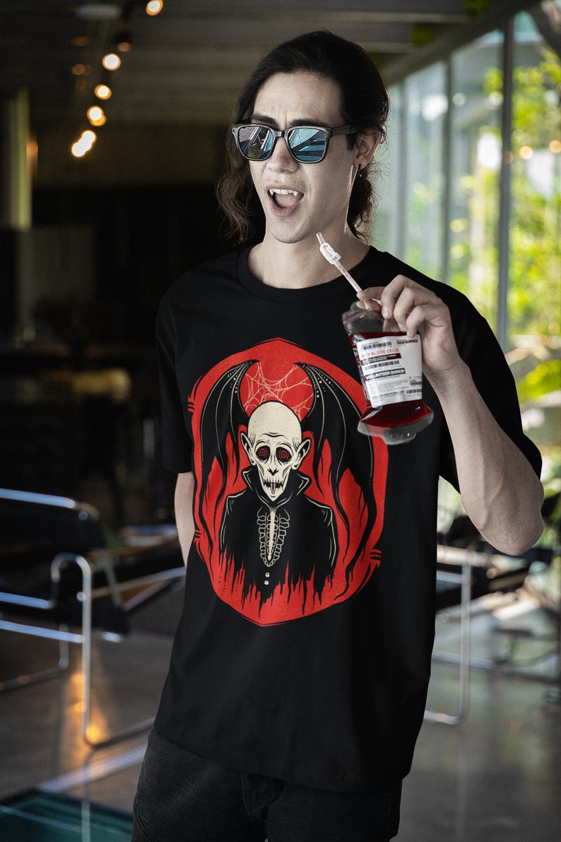 K&ouml;nnte beinhalten: Ein schwarzes T-Shirt mit einem Aufdruck eines Vampirs mit roten Augen und Fl&uuml;geln. Der Vampir tr&auml;gt einen schwarzen Anzug und hat ein wei&szlig;es, sch&auml;delartiges Gesicht. Der Aufdruck ist von einem roten Kreis mit Flammen umgeben. Die Person, die das Shirt tr&auml;gt, h&auml;lt einen Plastikbeutel mit roter Fl&uuml;ssigkeit und einem Strohhalm.