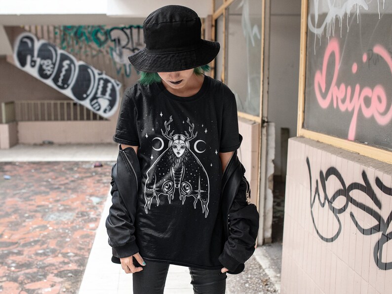 May include: A black t-shirt featuring a graphic print of a moth with a woman's face and antlers. The moth has crescent moons on its wings. The shirt is being worn by a person with green hair and a black hat.
