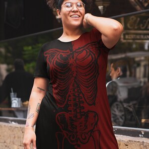 K&ouml;nnte beinhalten: Ein schwarzes und rotes T-Shirt-Kleid mit einem Skelett-Grafikdruck. Das Skelett ist in roter Farbe und auf der rechten Seite des Kleides gedruckt. Die linke Seite des Kleides ist schwarz.