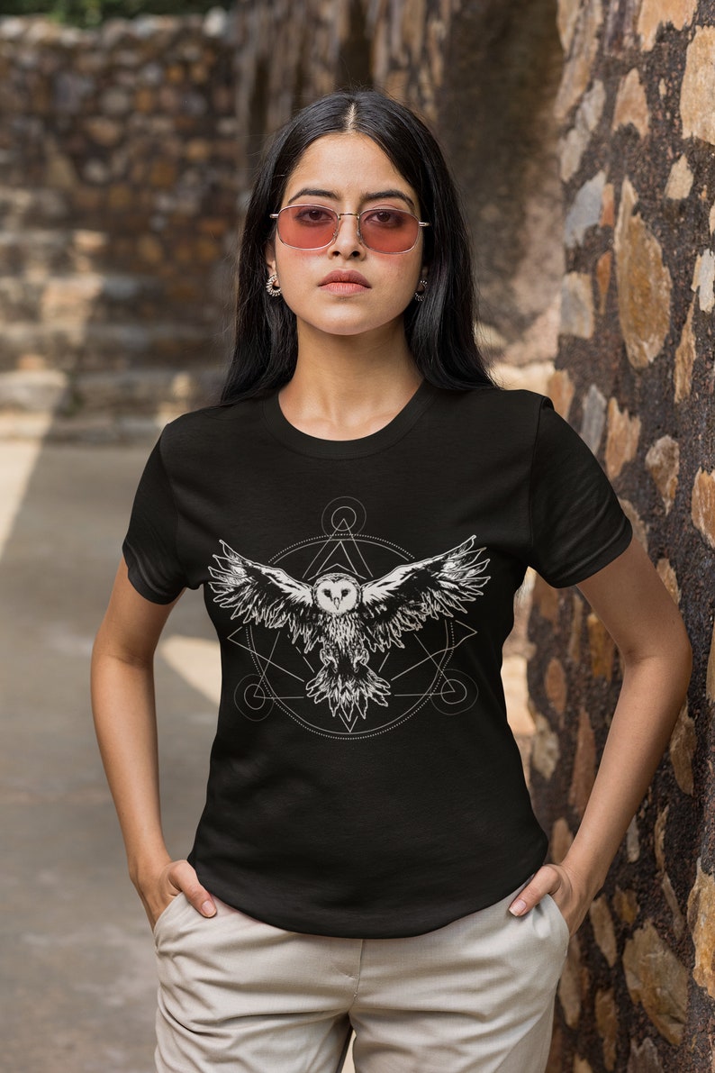 K&ouml;nnte beinhalten: Schwarzes T-Shirt mit einem wei&szlig;en Grafikdruck eines fliegenden Eulen innerhalb eines geometrischen Designs.