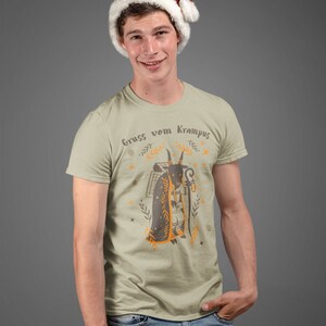 Puede incluir: Un hombre con una camiseta beige con un gr&aacute;fico de una figura de Krampus y el texto "Gruss vom Krampus". Tambi&eacute;n lleva una gorra de Pap&aacute; Noel roja y pantalones vaqueros azules.