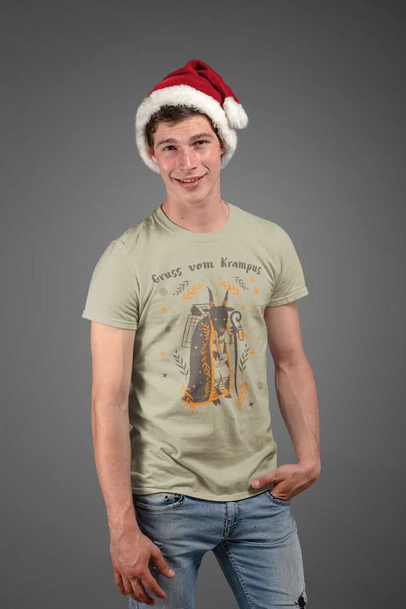 Puede incluir: Un hombre con una camiseta beige con un gr&aacute;fico de una figura de Krampus y el texto "Gruss vom Krampus". Tambi&eacute;n lleva una gorra de Pap&aacute; Noel roja y pantalones vaqueros azules.