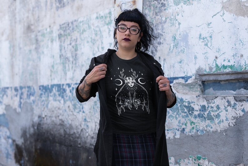 May include: Black t-shirt featuring a white graphic of a woman with antlers and a crescent moon on her head. The graphic is surrounded by stars.
