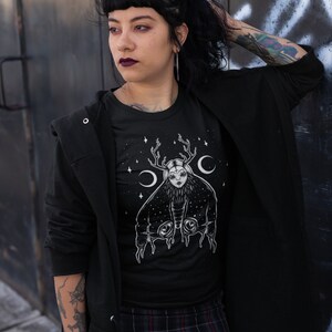 May include: A black t-shirt featuring a white graphic of a woman with antlers and wings. The graphic is surrounded by stars and crescent moons.