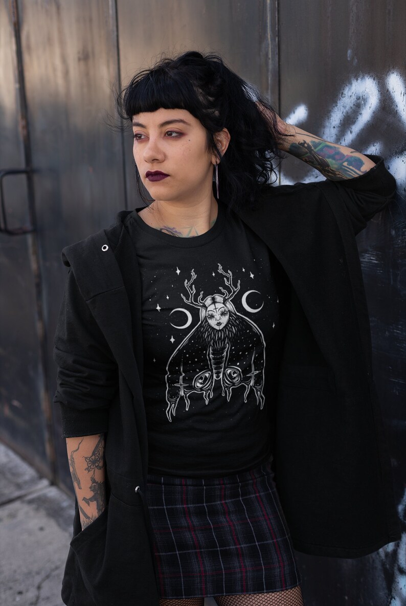 May include: A black t-shirt featuring a white graphic of a woman with antlers and wings. The graphic is surrounded by stars and crescent moons.