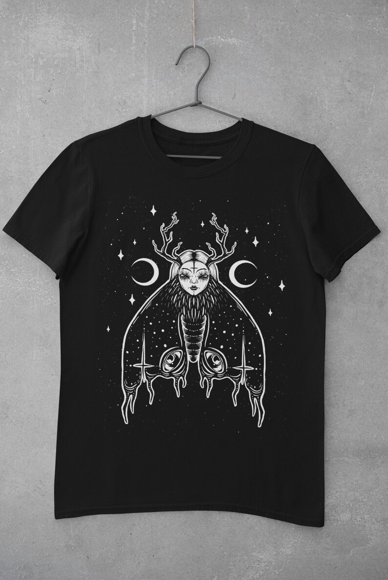May include: Black t-shirt featuring a white graphic of a moth with a human face and antlers. The moth is surrounded by stars and crescent moons.