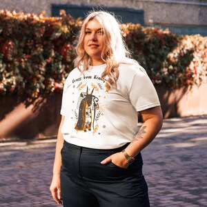 Puede incluir: Una camiseta blanca con una ilustraci&oacute;n en negro y naranja de una criatura con cuernos y un bast&oacute;n. El texto "Gruss vom Krampus" est&aacute; impreso en la camiseta.